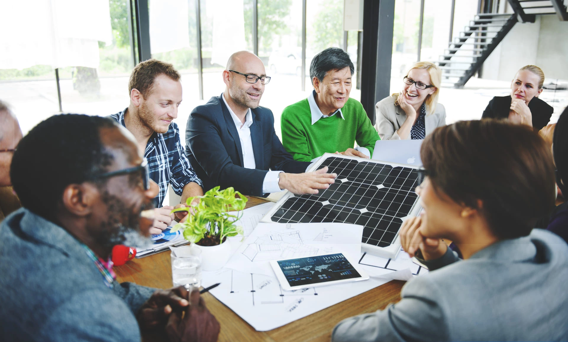 Gruppe von Geschäftsleuten bei einem Meeting, die gemeinsam über ein Solarpanel und technische Zeichnungen diskutieren – Symbol für nachhaltige Energie und Teamarbeit.