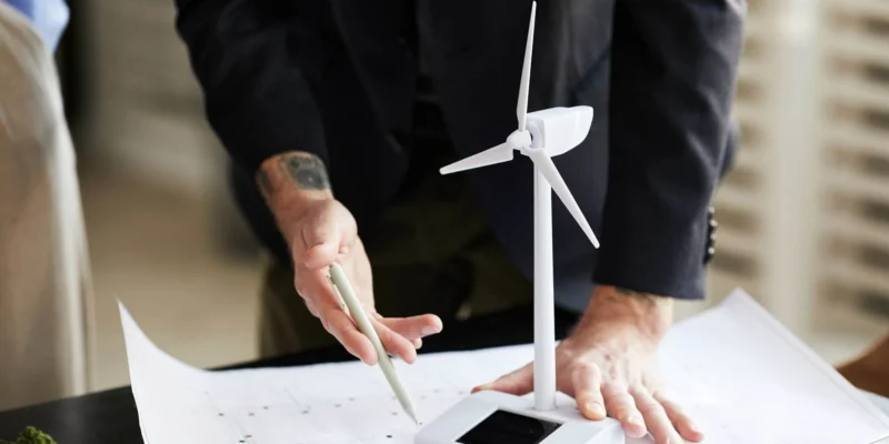 Eine Nahaufnahme von Händen, die über technischen Zeichnungen auf einem Tisch lehnen, mit einem kleinen Modell einer Windturbine im Fokus. Eine Person in einem dunklen Sakko hält einen Stift in der Hand, während eine andere Person teilweise im Bild links zu sehen ist. Es soll die Photovoltaik Förderung 2025 symbolisieren.