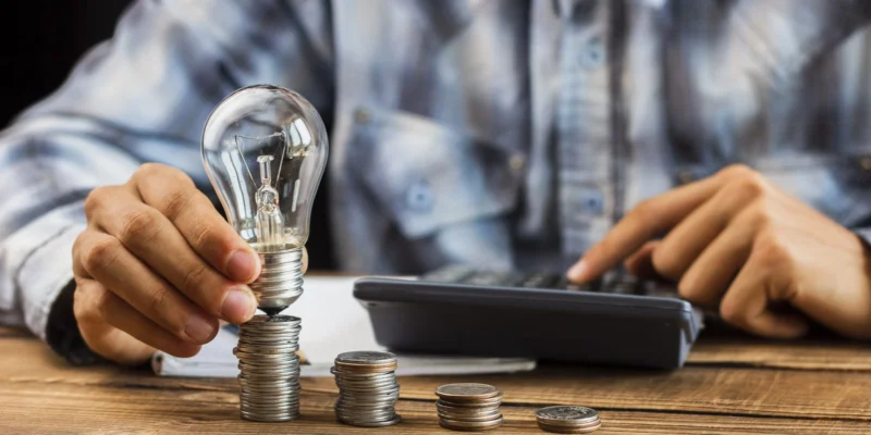 Mann in kariertem Hemd hält eine Glühbirne auf einem Stapel Münzen, im Hintergrund eine Hand auf einem Taschenrechner – Symbolbild für Energieeinsparung oder Investitionen in Ideen.