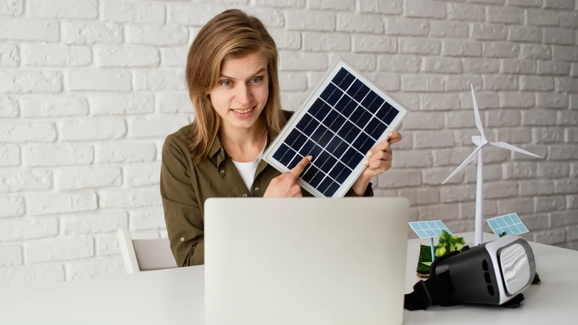 Eine junge Frau sitzt an einem Tisch und hält ein kleines Solarpanel in die Kamera, während sie vor einem Laptop sitzt. Im Hintergrund sind Modelle einer Windturbine und weiterer Solarpanels zu sehen. Diese Szene kann eine Präsentation oder Schulung über erneuerbare Energien und die damit verbundenen Steuervorteile durch Photovoltaikanlagen darstellen.