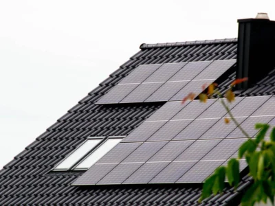 Auf einem dunkelgrauen, geneigten Dach mit schwarzen Dachziegeln sind rechteckige Solarpanels angebracht. Das Dach hat außerdem zwei rechteckige Dachfenster. Ein schwarzer Schornstein ist im oberen Bereich des Daches zu sehen. Im Vordergrund ist ein unscharfer grüner Zweig zu erkennen.