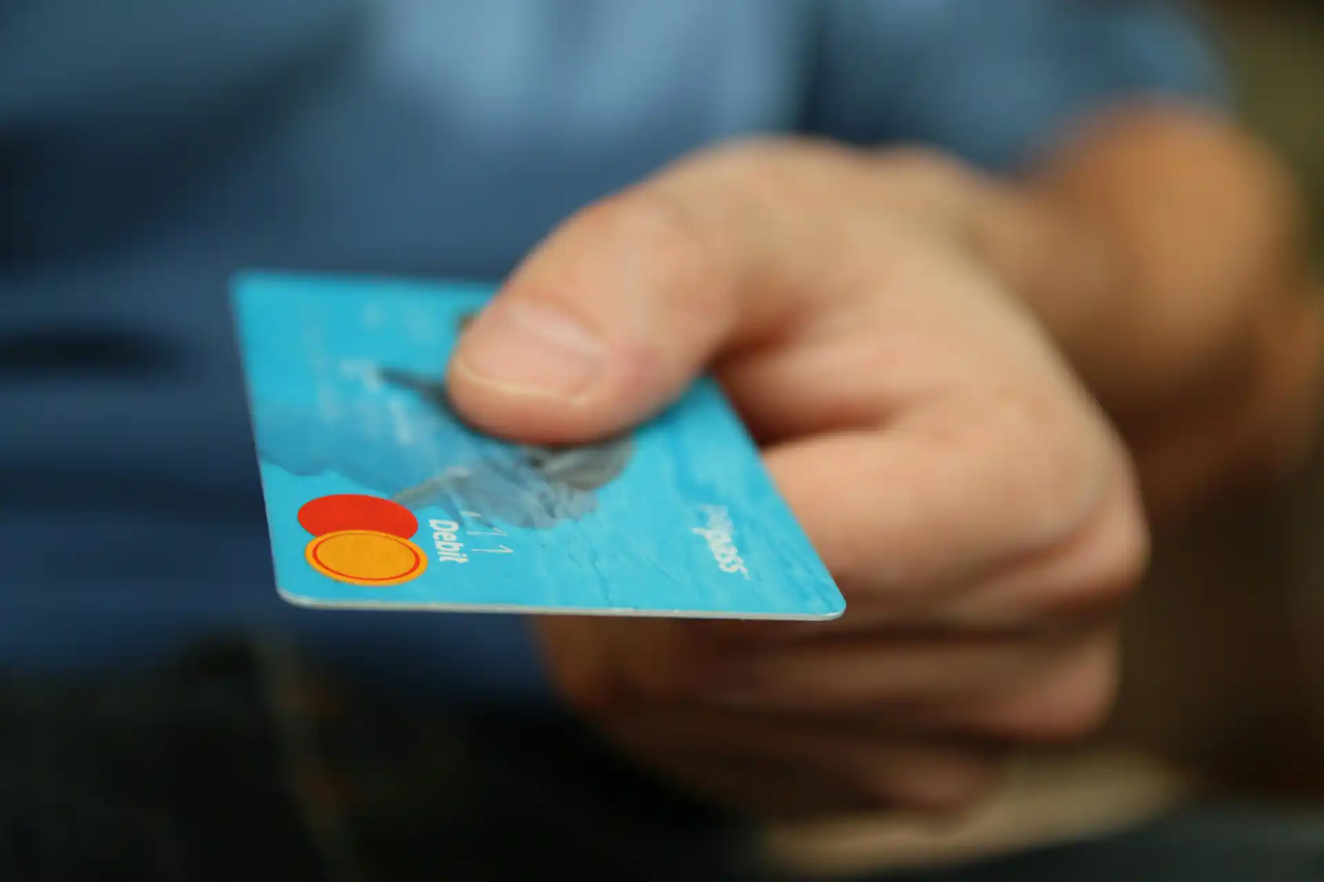 Nahaufnahme einer Hand, die eine blaue Debitkarte mit Mastercard-Logo zum Bezahlen überreicht – Symbolbild für bargeldlosen Zahlungsverkehr.