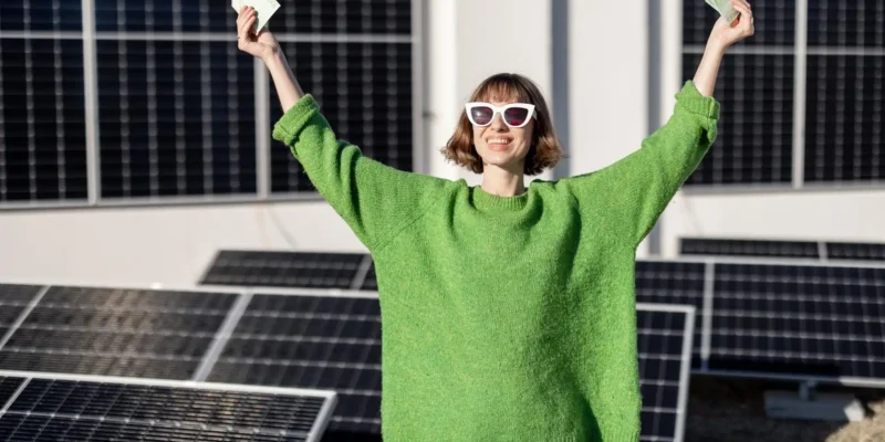 Eine fröhliche Frau steht vor einer Anlage mit Solarpanels, trägt einen leuchtend grünen Pullover und weiße Sonnenbrille. Sie hält in beiden Händen Geldscheine hoch und lächelt strahlend, was Freude und die Kosten und Ersparnis durch Photovoltaikanlagen symbolisiert.