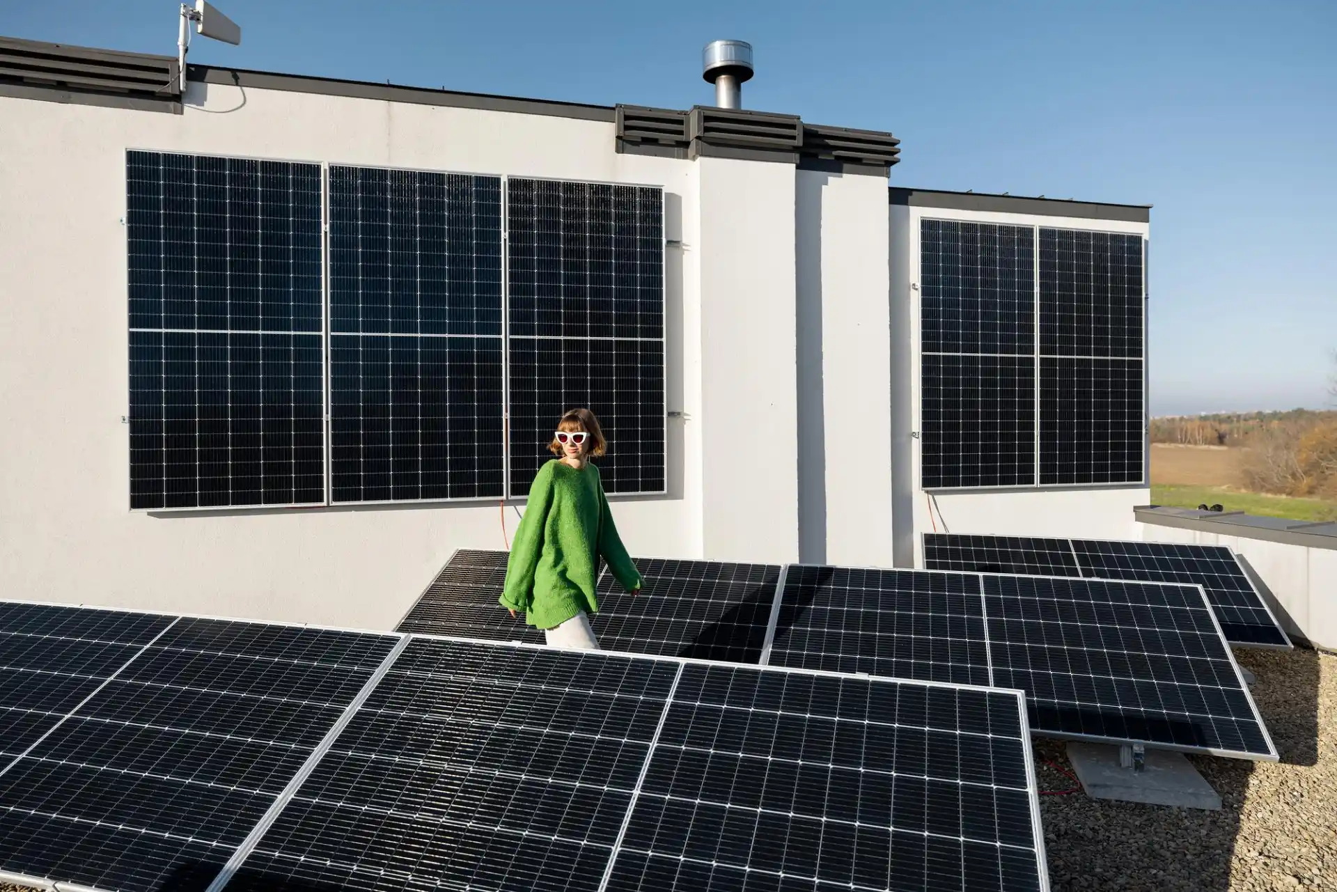 Eine Frau mit grünem Pullover und Sonnenbrille geht zwischen aufgestellten Solarpanels auf einem Flachdach entlang, im Hintergrund sind weitere Module an der Fassade montiert – das Bild veranschaulicht das Thema Photovoltaikanlagen Kosten und Ersparnis, indem es den Ausbau von Solartechnik zur Energiegewinnung und möglichen finanziellen Vorteilen zeigt.