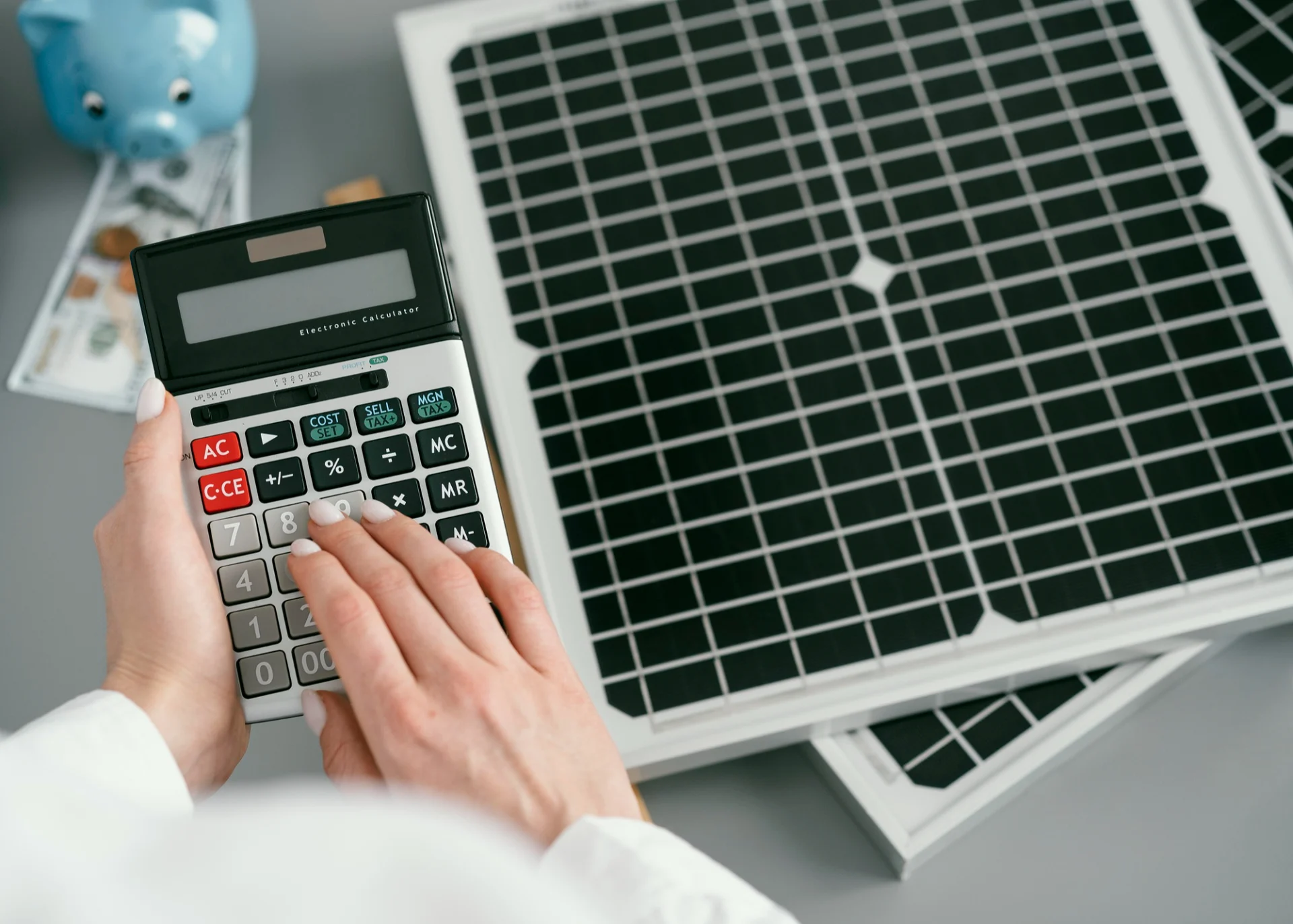 Nahaufnahme von Händen, die einen Taschenrechner benutzen, der auf einem Tisch neben einem Solarmodul liegt. Im unscharfen Hintergrund stehen ein blaues Sparschwein und Dollarnoten als Symbol für die Finanzierung oder Ersparnis durch Solarenergie.