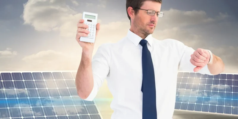 Geschäftsmann mit Brille hält einen Taschenrechner hoch und schaut auf seine Armbanduhr, während er vor zwei Solarmodulen und einem bewölkten Himmel steht – Symbol für die Kalkulation von Solarenergie.