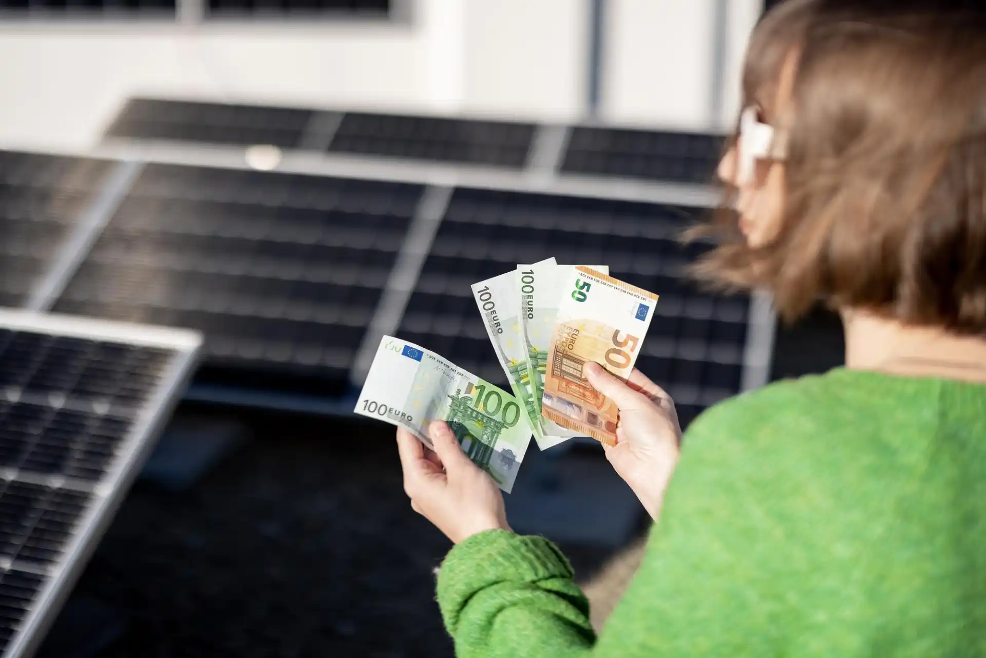 Eine Frau in grünem Pullover zählt mehrere Euro-Geldscheine vor einer Reihe von Solarpanels – das Bild symbolisiert das Thema Photovoltaikanlagen Kosten und Ersparnis, indem es aufzeigt, wie durch Solartechnik finanzielle Vorteile erzielt werden können.