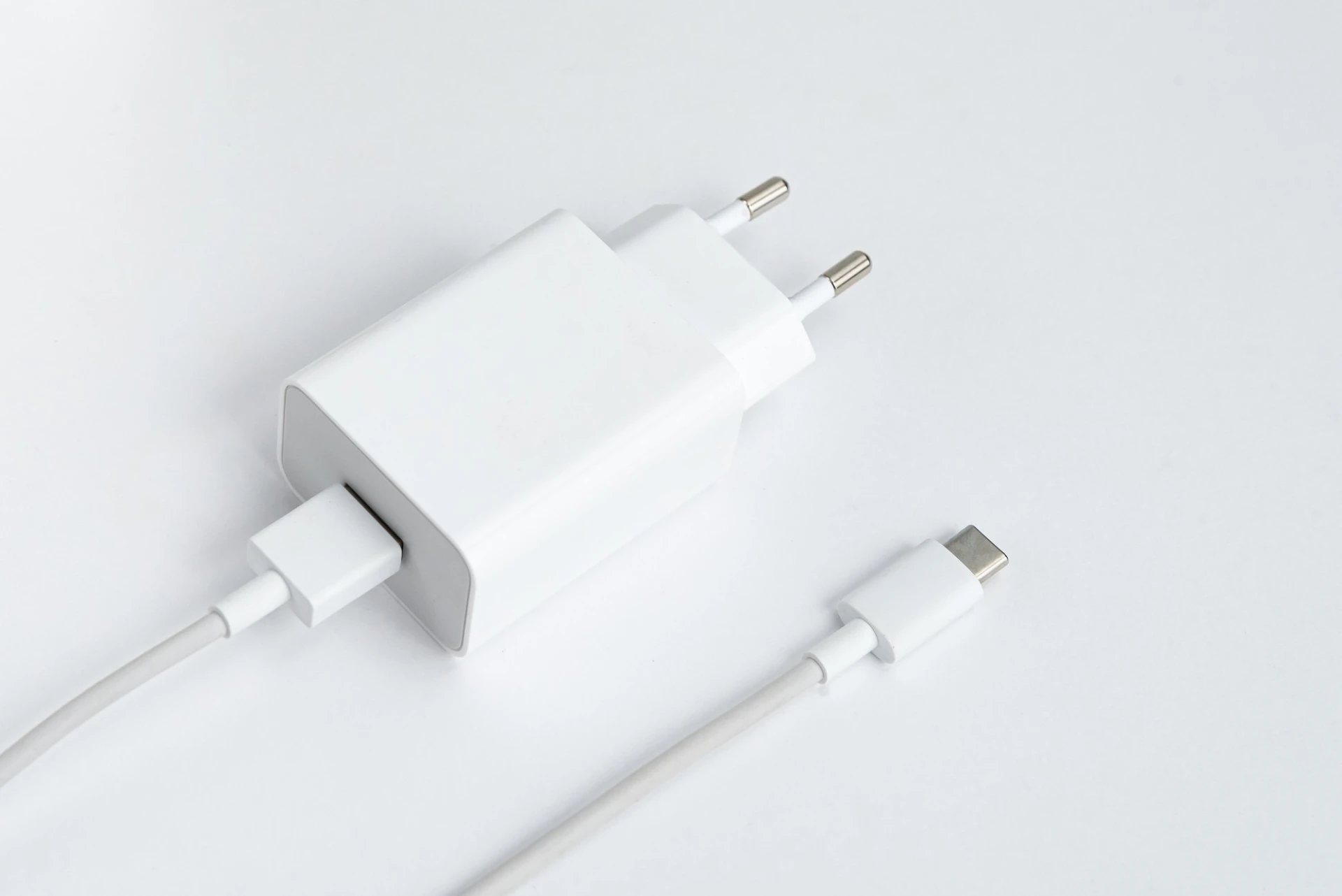 Ein horizontales Close-up zeigt ein weißes USB-Ladegerät und ein separates, hellgraues USB-C-Kabel auf einer glänzend weißen Oberfläche. Das Ladegerät hat einen europäischen Stecker (Schuko-ähnlich) und einen einzelnen USB-A-Anschluss, in den das USB-C-Kabel eingesteckt ist. Das Kabel verläuft diagonal über den unteren Bildbereich und endet in einem USB-C-Stecker. Die schlichte Darstellung und die helle Farbgebung lenken den Fokus auf die Funktionalität der Steckverbindung für die Stromversorgung moderner Geräte.