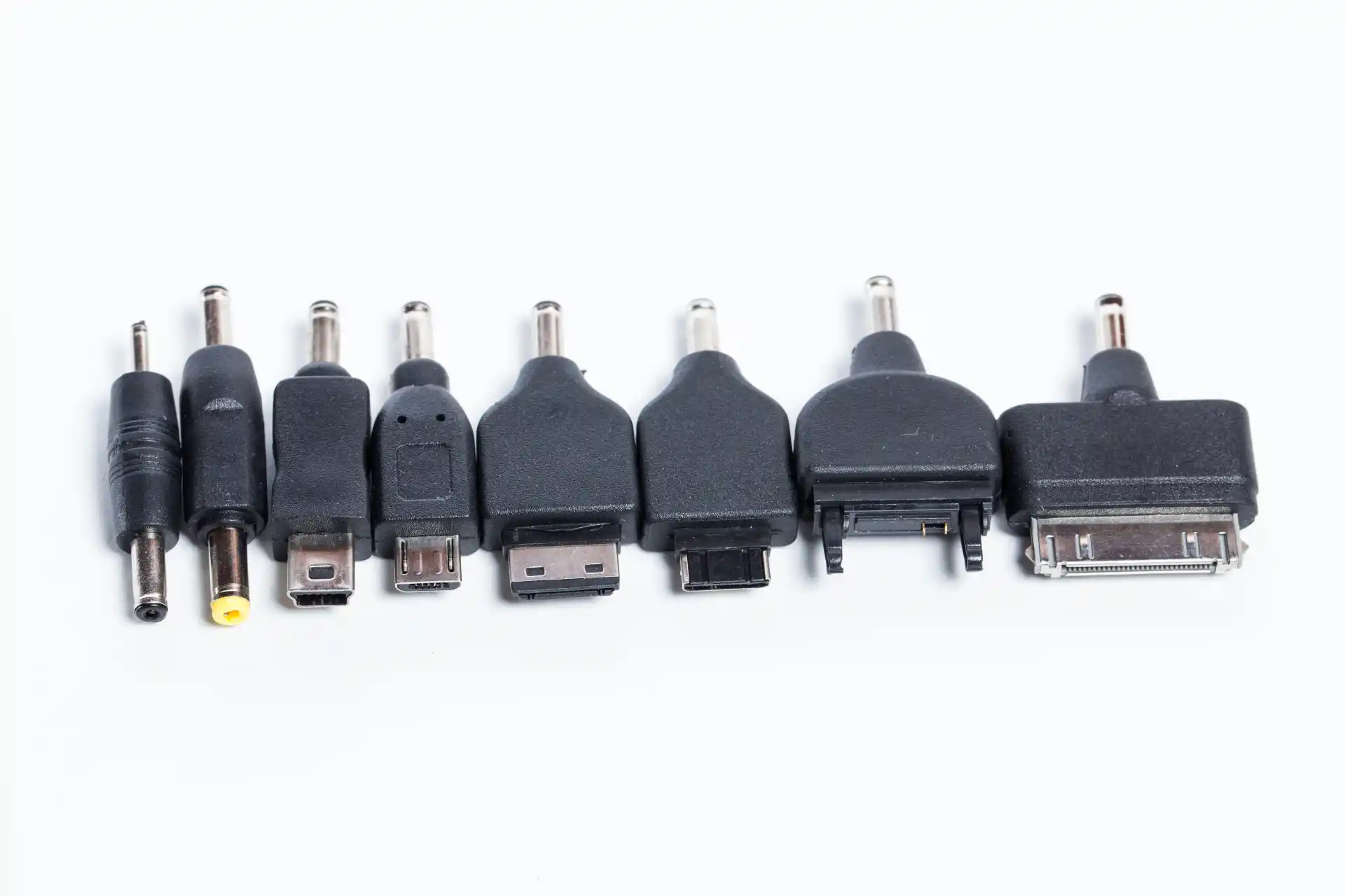 Verschiedene Adapteraufsätze für Ladegeräte von Mobiltelefonen und Elektronikgeräten in einer Reihe auf weißem Hintergrund, darunter USB, Mini-USB, Micro-USB und ältere DC-Stecker.