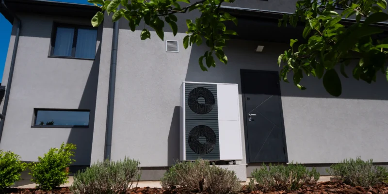 Das Bild zeigt eine Wärmepumpe, montiert an einer Hauswand neben einer Tür. Im Vordergrund ist die grüne Bepflanzung eines Vorgartens zu sehen.