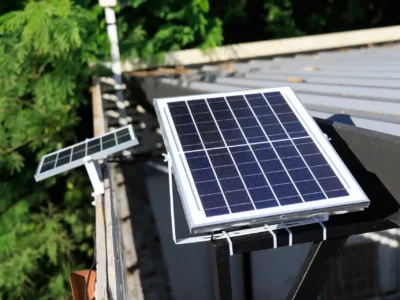 Nahaufnahme zweier kleiner Solarpanels, die auf einem Dach installiert sind. Die Paneele sind in Richtung Sonne ausgerichtet, im Hintergrund sind grüne Bäume sichtbar.