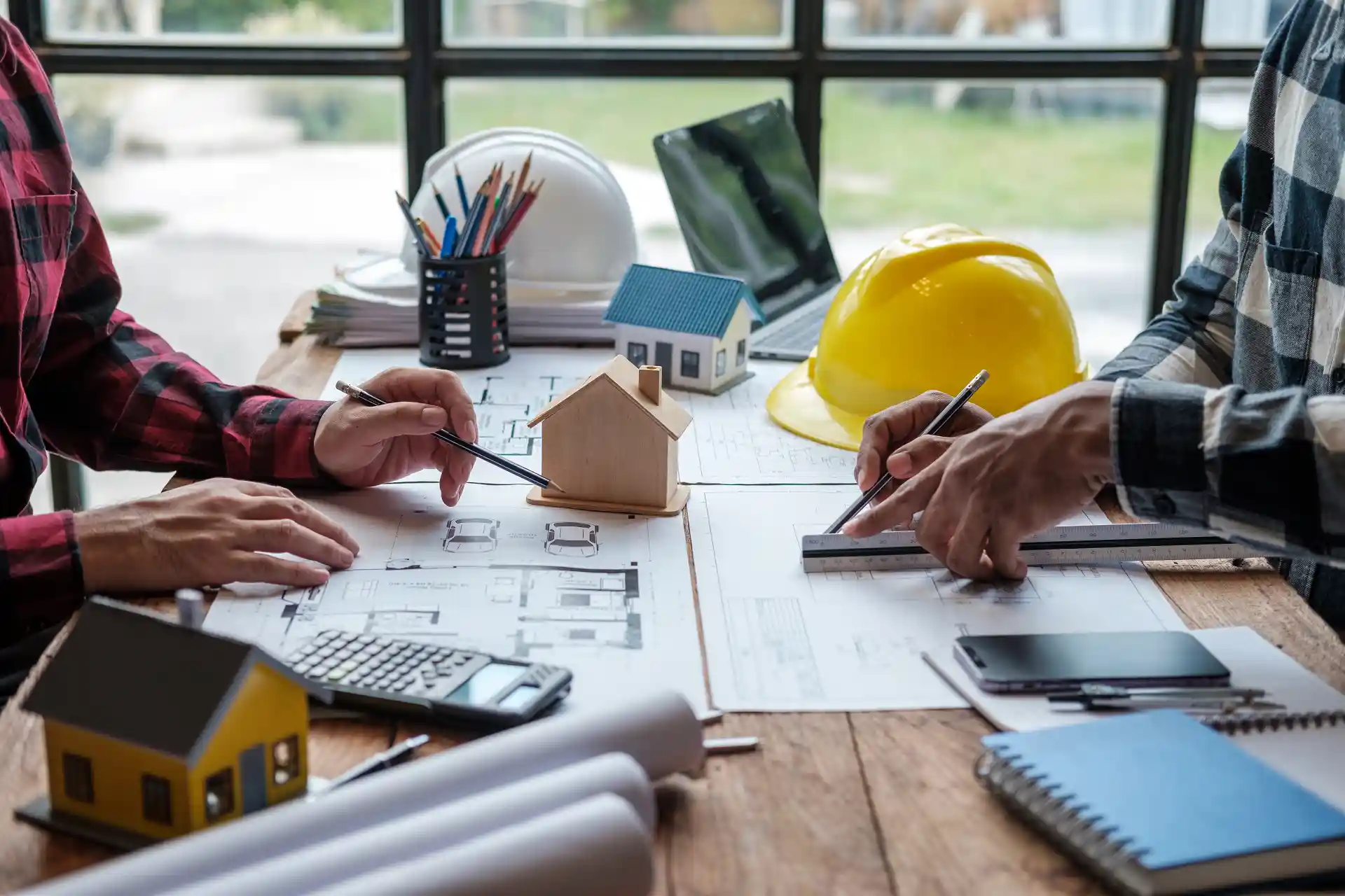 Ingenieure arbeiten an Bauplänen mit Modellhäusern und Schutzhelmen – Planung und Contracting im Bauwesen