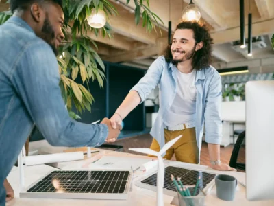 Zwei Männer schütteln sich in einem modernen Büro die Hände, wobei der eine eine Jeansjacke und der andere ein hellblaues Hemd und eine gelbe Hose trägt. Auf dem Tisch vor ihnen liegen Miniatur-Solarmodule und Windräder, die saubere Energie und grüne Technologie symbolisieren. Das Lächeln der Männer deutet auf eine erfolgreiche Zusammenarbeit oder einen Geschäftsabschluss im Bereich erneuerbarer Energien hin, möglicherweise im Kontext von Projekten zur Volleinspeisung von Solarstrom.