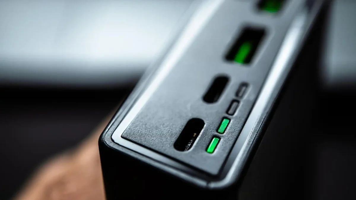 Ein Detail-Foto aus geringer Entfernung zeigt die Oberseite eines dunklen, rechteckigen elektronischen Geräts, bei dem es sich um einen Laderegler oder eine Powerbank handeln könnte. Der Fokus liegt auf den Anschlüssen und der Statusanzeige an der Vorderkante. Drei kleine, rechteckige LEDs leuchten grün und zeigen wahrscheinlich den Ladestatus oder die Betriebsfunktion des Geräts an. Das Bild symbolisiert den technischen Aspekt der Ladesteuerung.